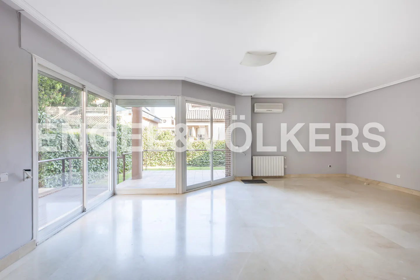 Bright, empty room with marble floors, gray walls, and large windows overlooking a green yard. Radiator and AC unit visible.