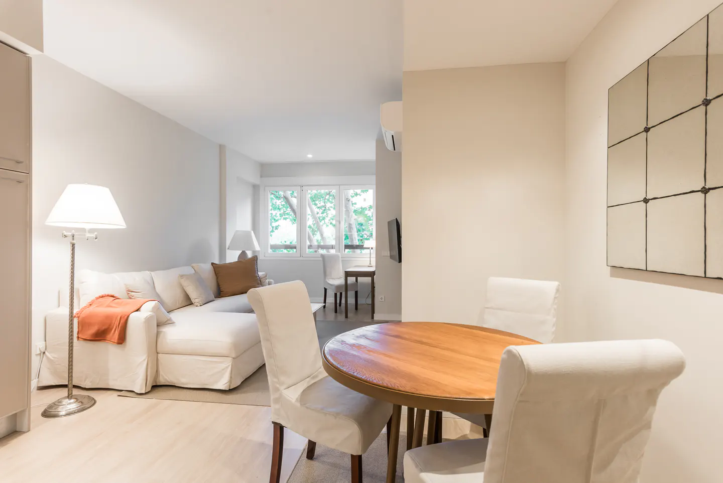 Living room with a round wooden table, white chairs, and a white sofa with pillows and a blanket. A grid mirror hangs on the wall.