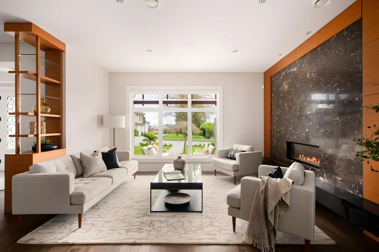 Bright living room with a gray sofa, two armchairs, and a black coffee table on a patterned rug. A fireplace and large window offer views of the green lawn.