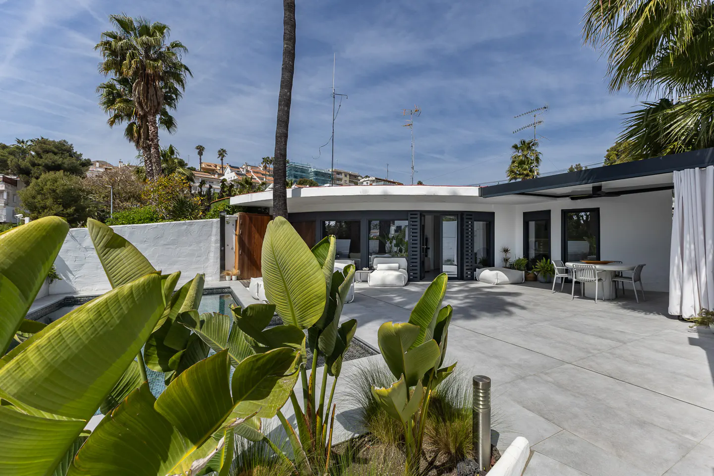 Modern white house with a gray patio, table, chairs, and white cushions. Tropical plants and palm trees surround the property.