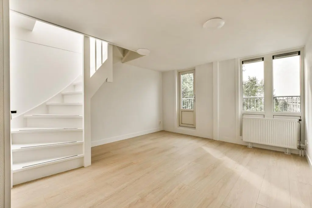 Bright, minimalist room with light wood floors, white walls, a white staircase, and windows overlooking a balcony.
