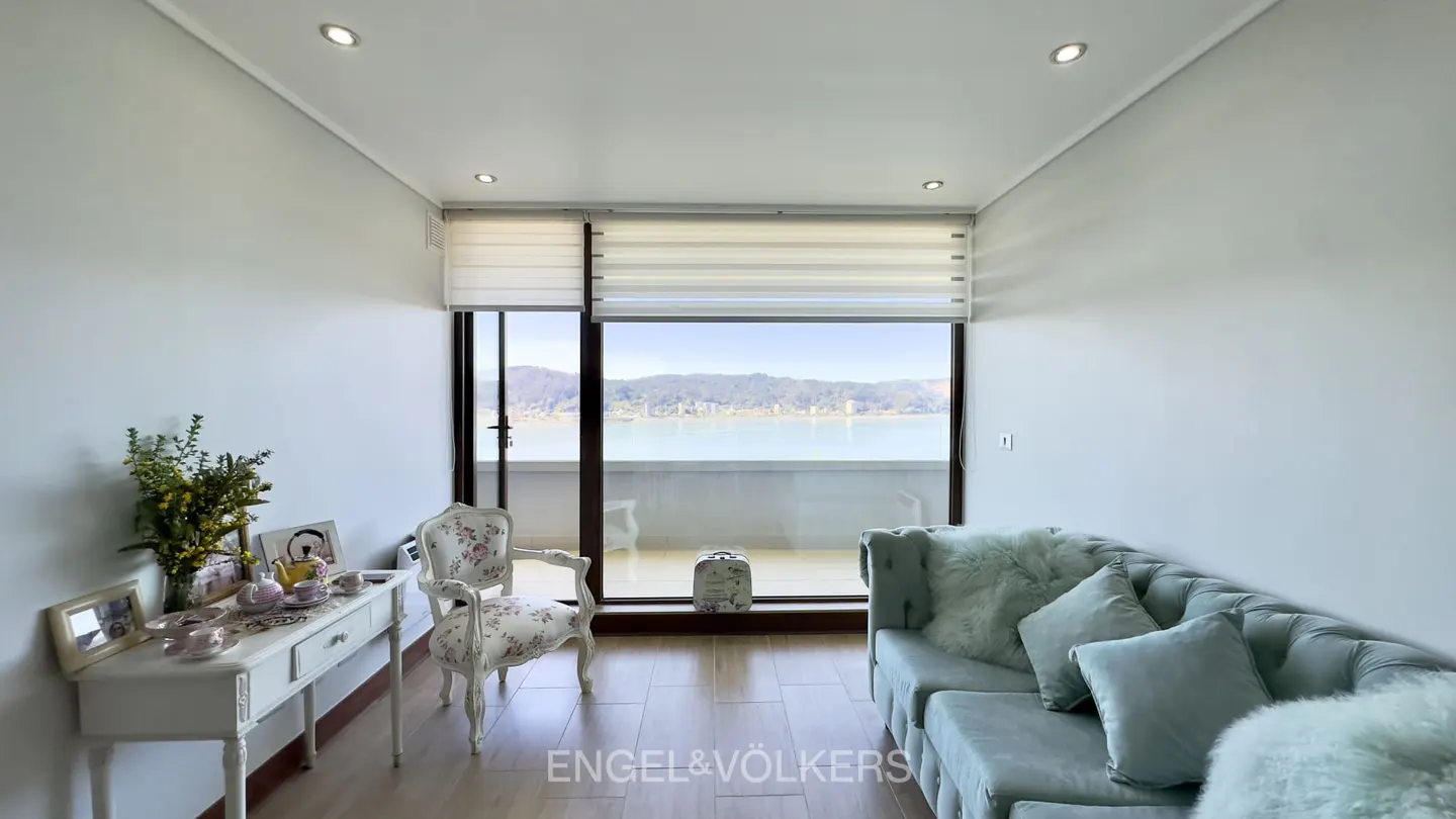 Bright living room with a light blue tufted sofa, floral armchair, white console table, and a balcony view of the ocean.