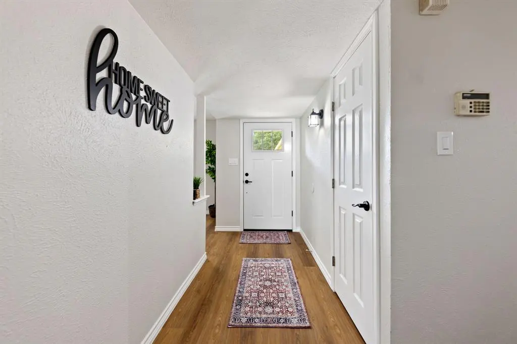 Hallway with wood floors, white walls, and a white front door. A black "Home Sweet Home" sign hangs on the wall. Two rugs line the floor.