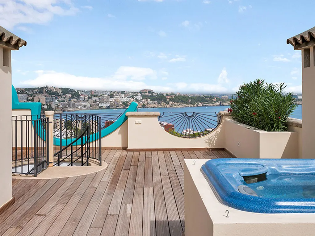 Rooftop deck with a blue hot tub, wooden flooring, and a view of the ocean and city skyline under a blue sky.