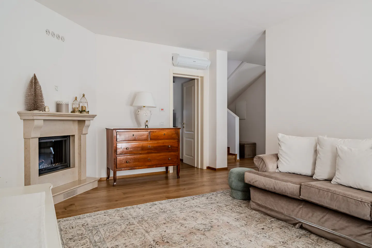Living room with a beige sofa, white pillows, a fireplace, and a wooden dresser on a patterned rug.