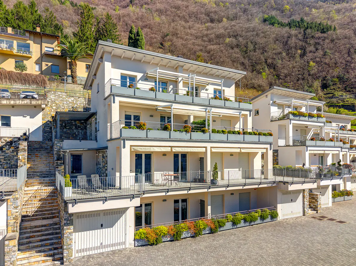 Modern white apartment building with balconies and flower boxes, set against a hillside with trees. Garage doors are visible on the ground floor.