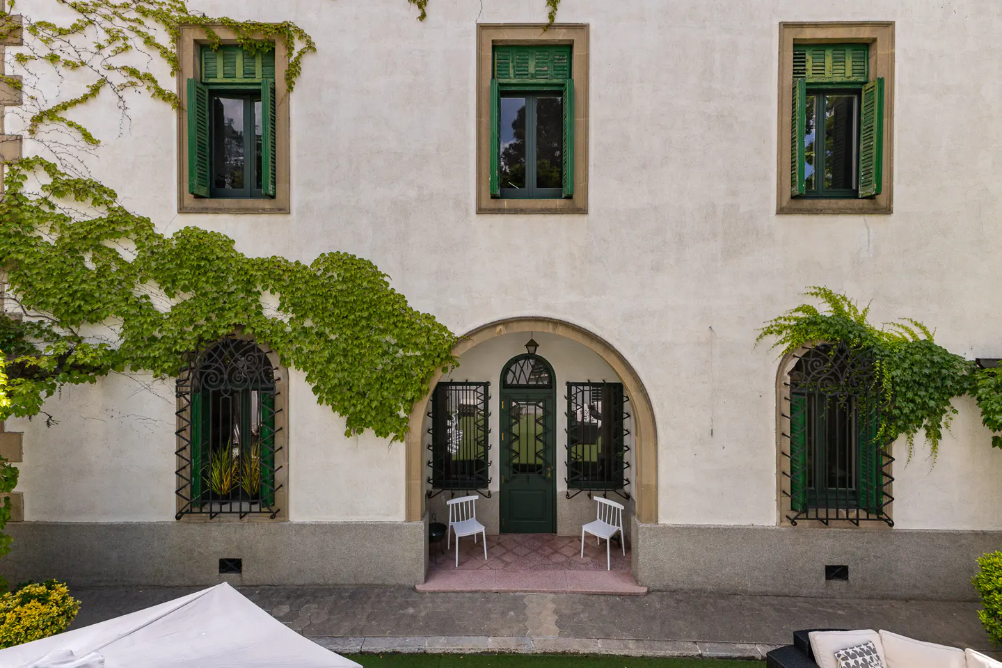 Exterior of a white building with green shutters and ivy. An arched doorway has a green door and two white chairs.