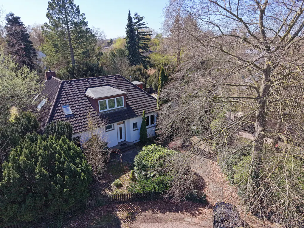 Aerial view of a white house with a brown tiled roof, surrounded by lush green trees and a brick driveway.