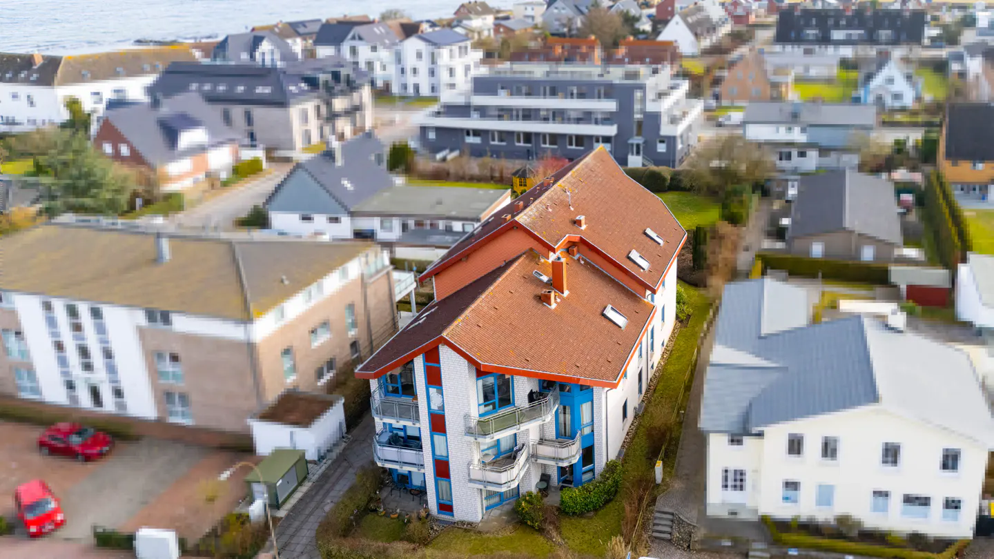 Aerial view of a three-story building with a red-tiled roof and blue accents, surrounded by other houses in a coastal town.
