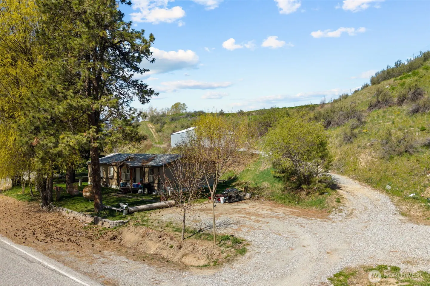 A landscape view of a small, rustic house with a gravel driveway and a grassy hillside backdrop.