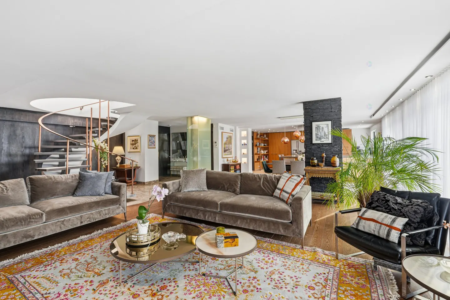 A spacious living room with two brown sofas, a black chair, and a colorful rug. A spiral staircase is visible in the background.