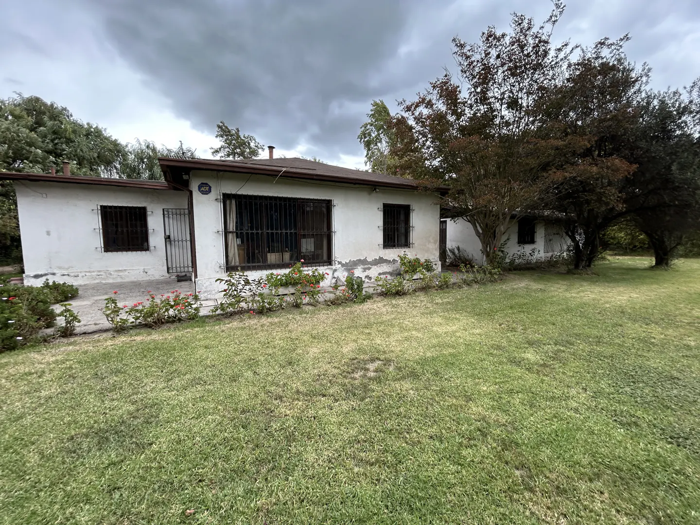 Exterior view of a one-story white house with bars on the windows, surrounded by a green lawn and trees under a cloudy sky.