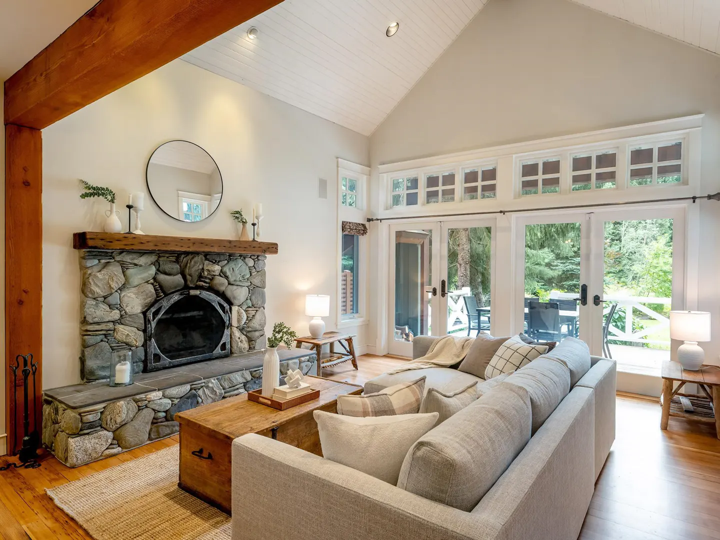 Bright living room with stone fireplace, wood beam, and vaulted ceiling. A gray sectional faces French doors to a deck.