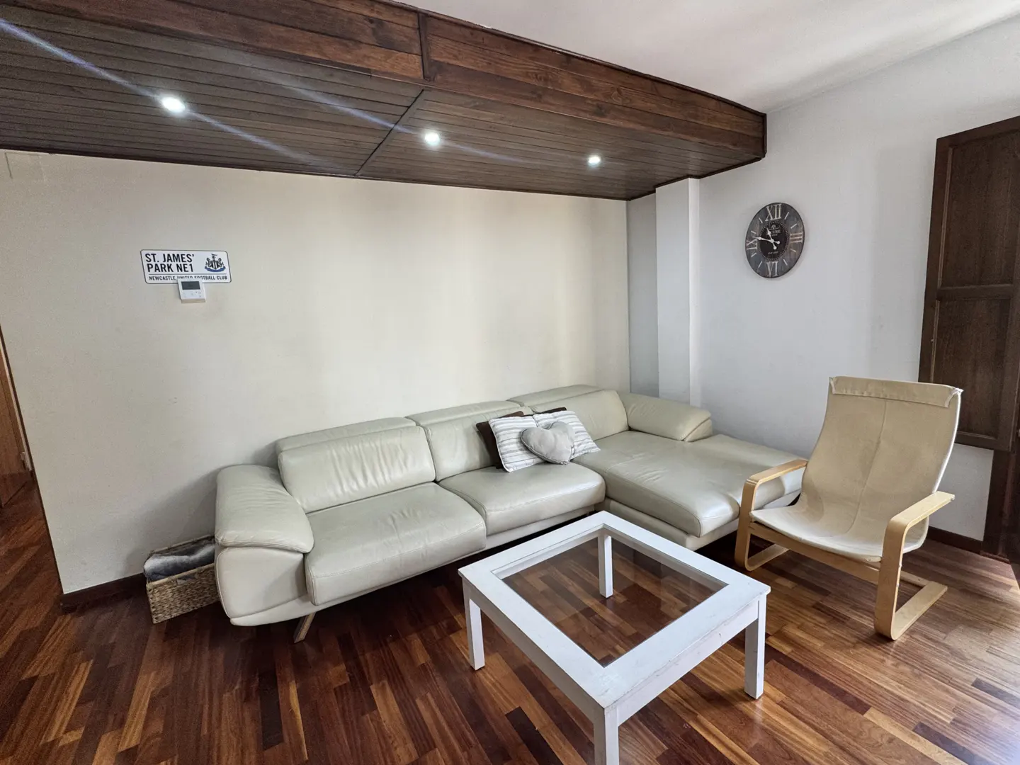 Living room with a white leather sectional sofa, white coffee table, and a beige armchair on a wood floor. A clock hangs on the wall.