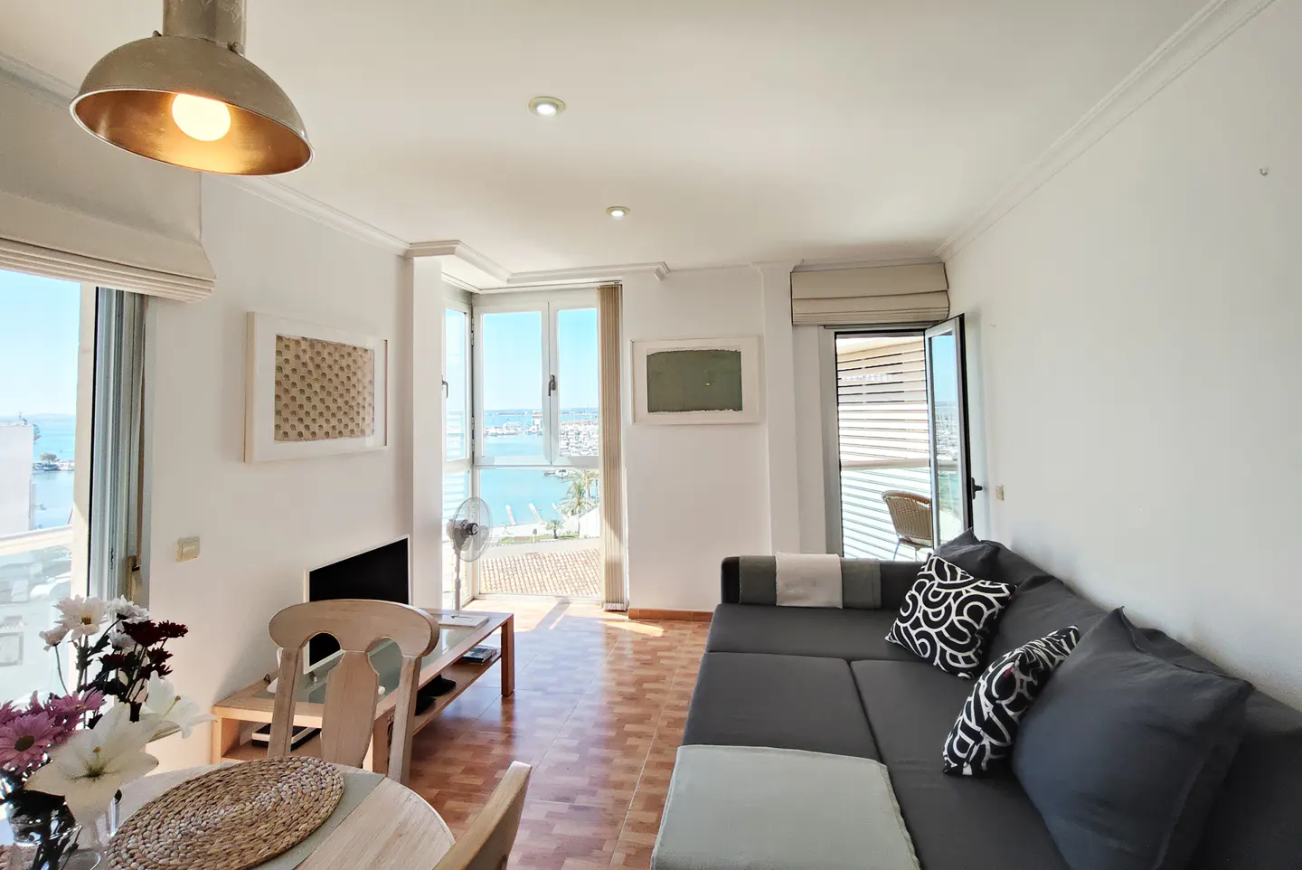Bright living room with a gray sectional sofa, wood floors, and a view of the ocean through open windows.