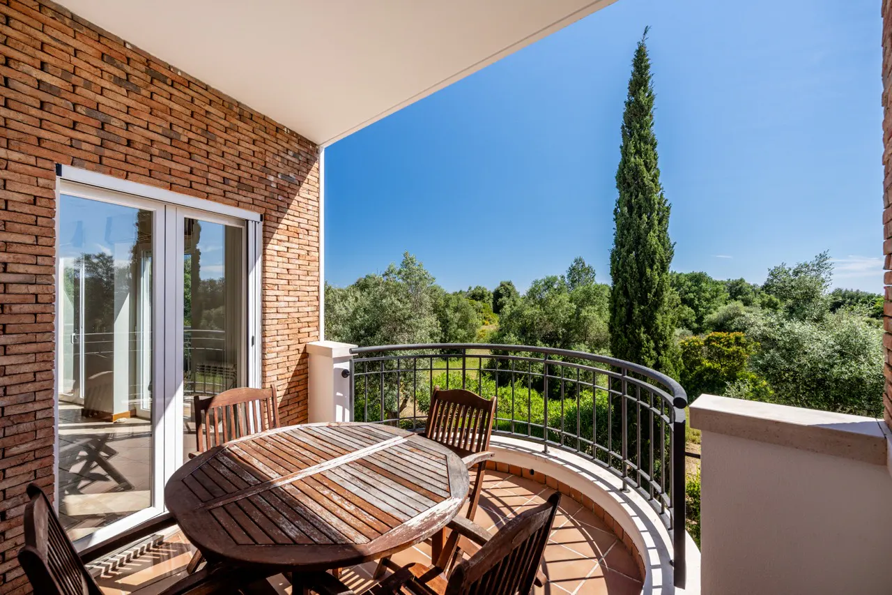 Balcony with a round wooden table and chairs, overlooking green trees and a blue sky. The balcony has a brick wall and a black metal railing.