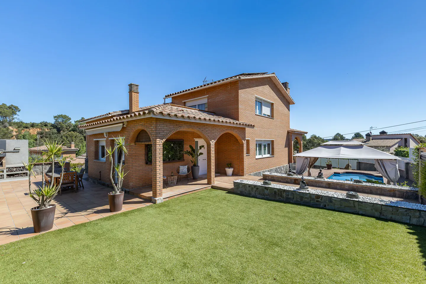 Two-story brick house with arched porch, green lawn, and pool with gazebo on a sunny day.