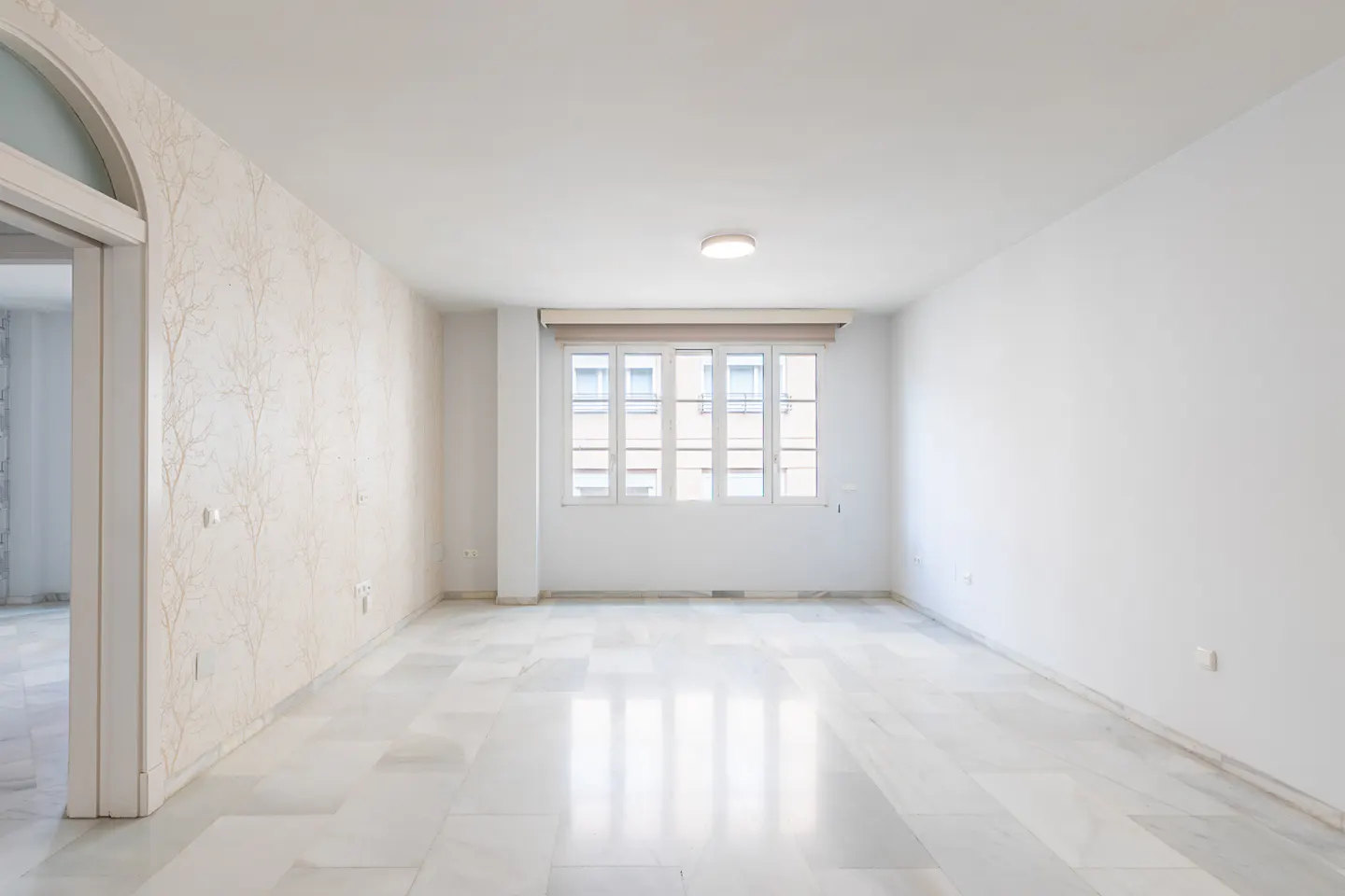 Bright, empty room with white walls, marble floors, and a large window. A doorway is visible on the left.