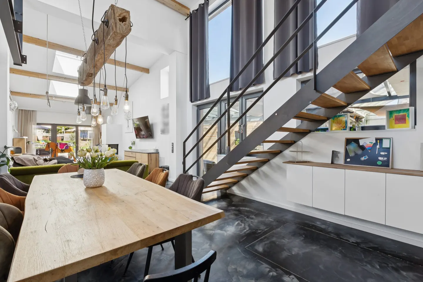 Bright, modern interior with a wooden dining table, black chairs, and a staircase with metal railings. White walls and a large window add to the airy feel.