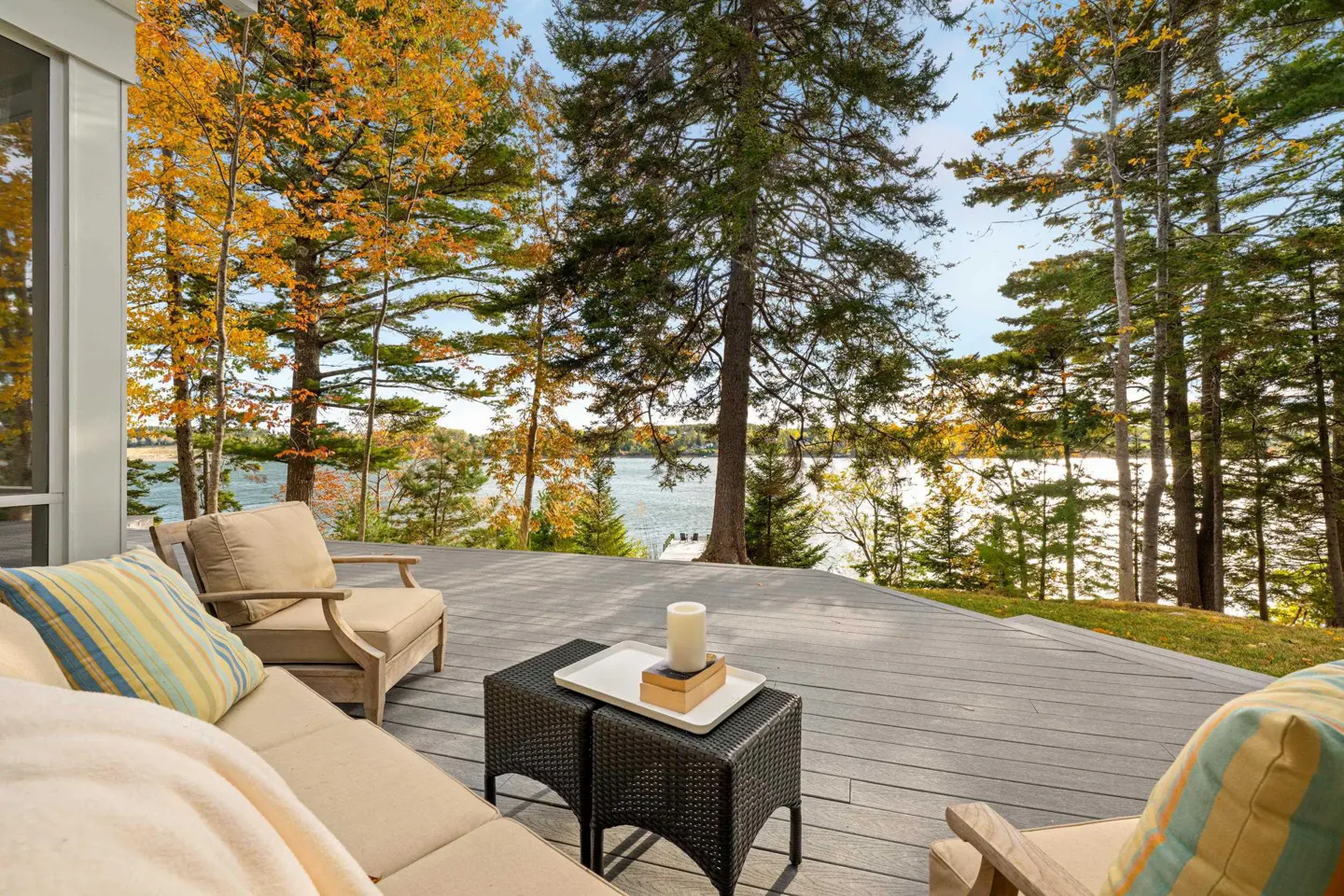Outdoor deck with wicker table, beige cushions, and striped pillows overlooking a lake surrounded by trees in autumn colors.