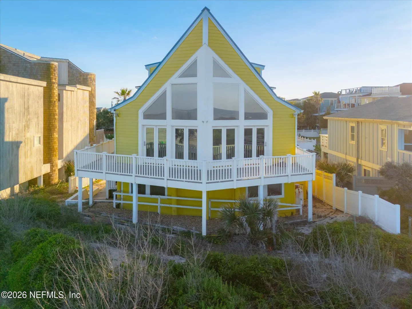 Bright yellow A-frame beach house with a large white deck and expansive windows, surrounded by coastal dunes and greenery.