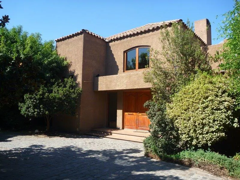 Exterior view of a two-story stucco house with a red tile roof and a wooden double door entrance. Green trees surround the house.