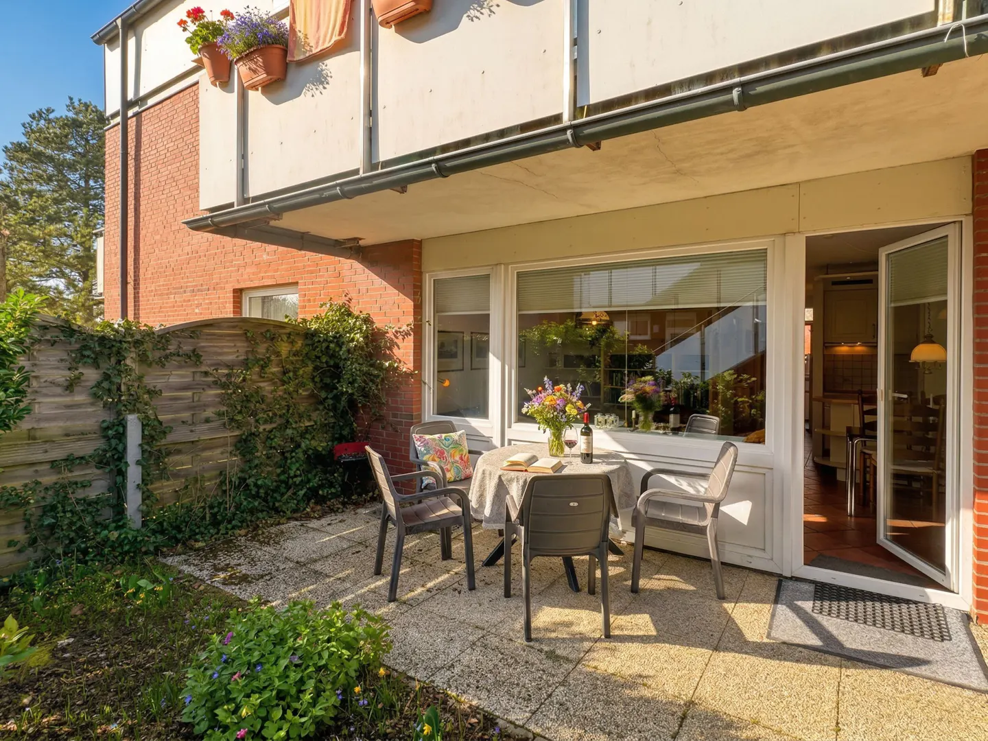 Outdoor patio with table, chairs, flowers, and wine. Brick building with balcony and flower boxes in the background.