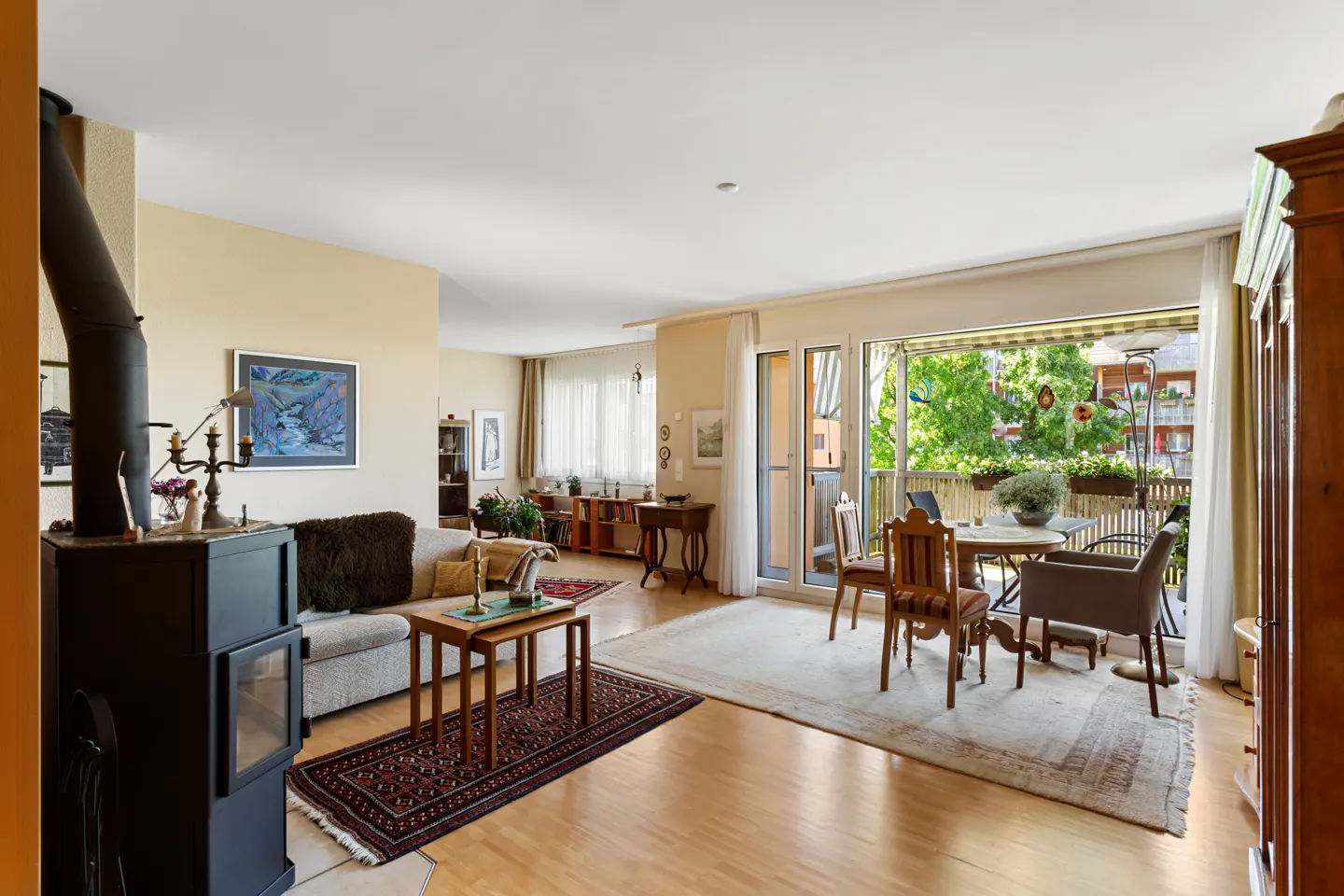 Bright living room with wood floors, a black fireplace, a gray sofa, and a dining table on a balcony with green trees.