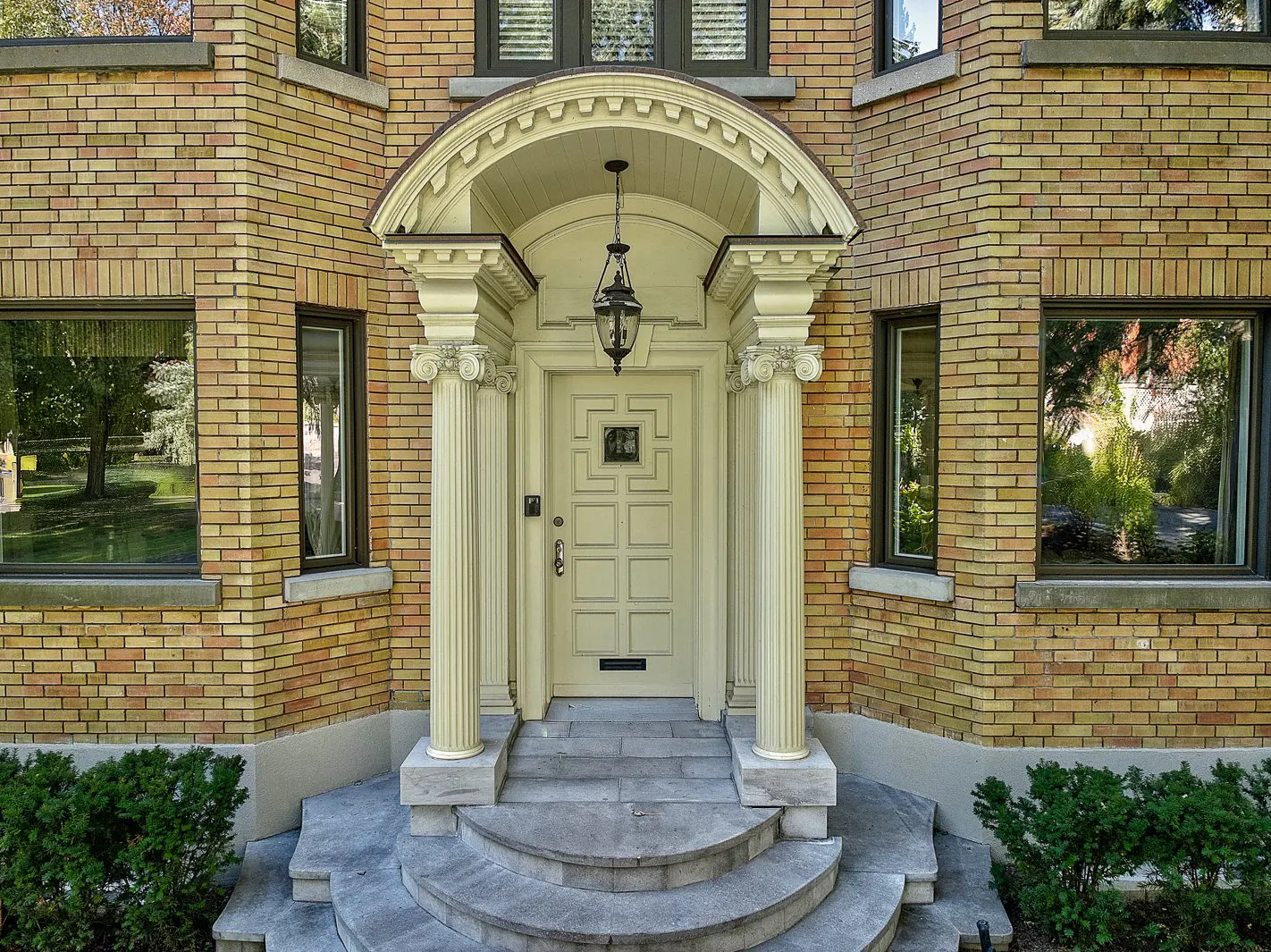Front view of a brick house with a cream-colored door framed by columns and a curved archway. A hanging lantern illuminates the entrance.