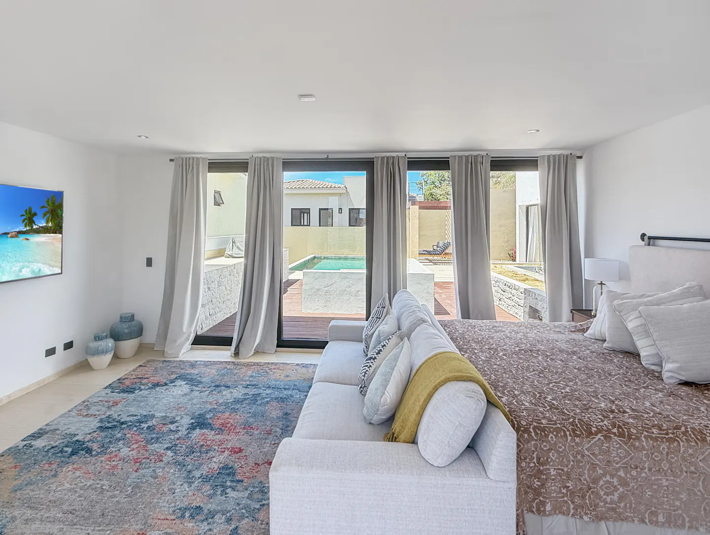 Bright bedroom with a sofa at the foot of the bed, a colorful rug, and a view of the pool through sliding glass doors.