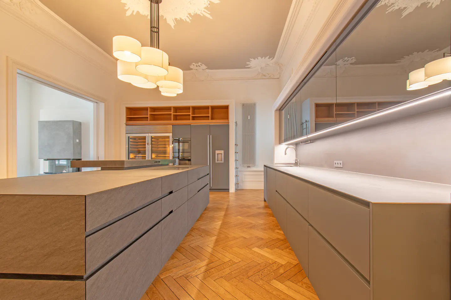 A modern kitchen with gray cabinets, a large island, and herringbone wood floors. A chandelier hangs from the ornate ceiling.