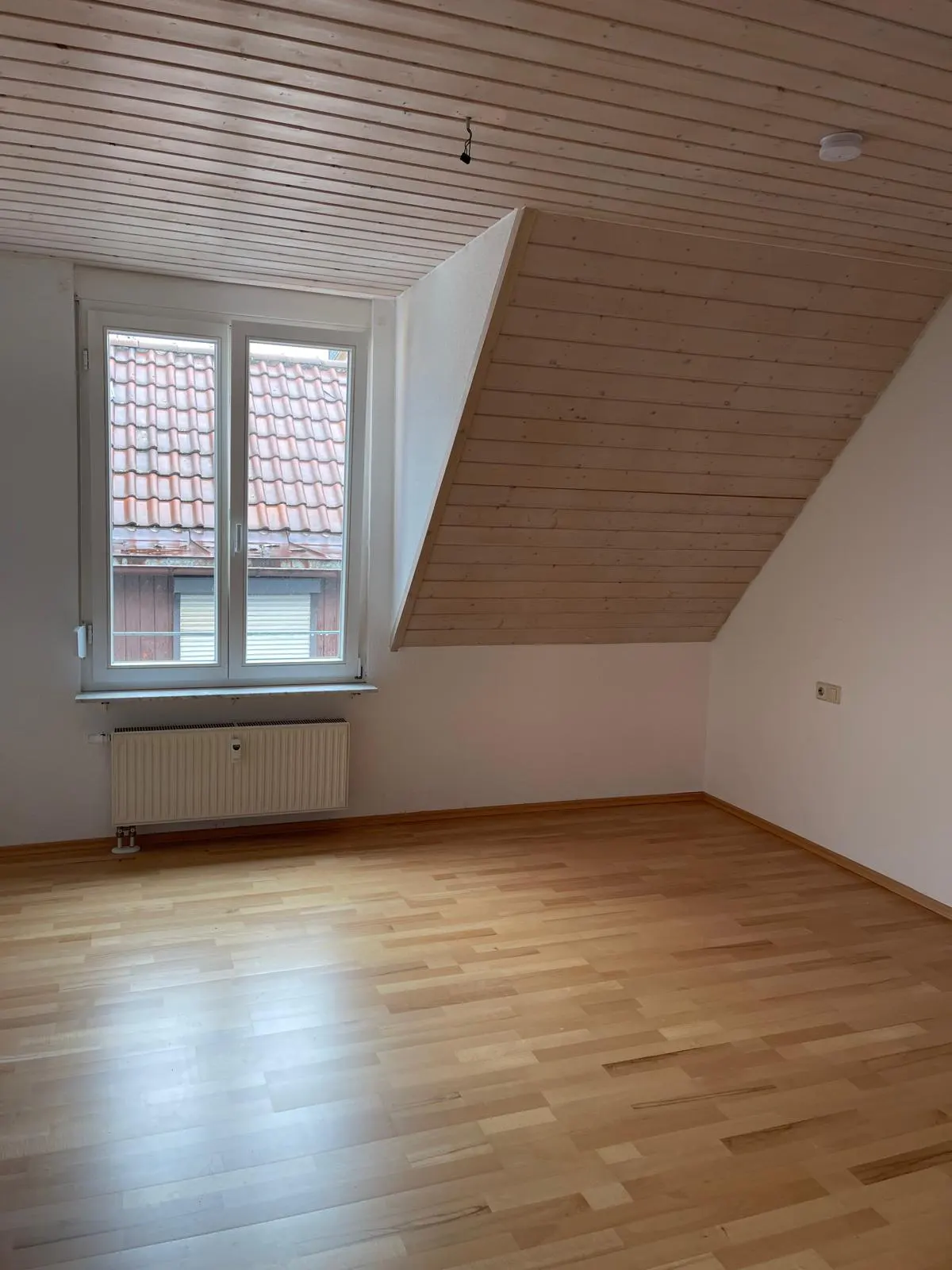 Empty attic room with light wood flooring, sloped wood-paneled ceiling, white walls, and a window overlooking a roof.