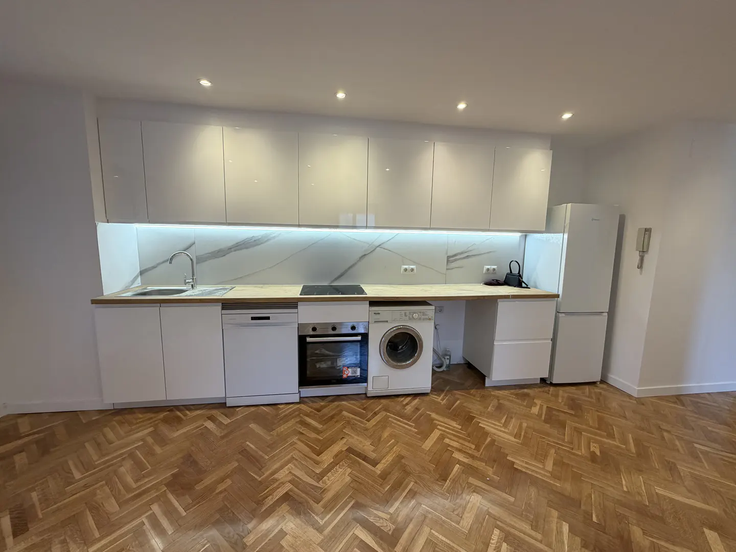 Modern white kitchen with marble backsplash, wooden countertops, built-in appliances, and herringbone wood flooring.