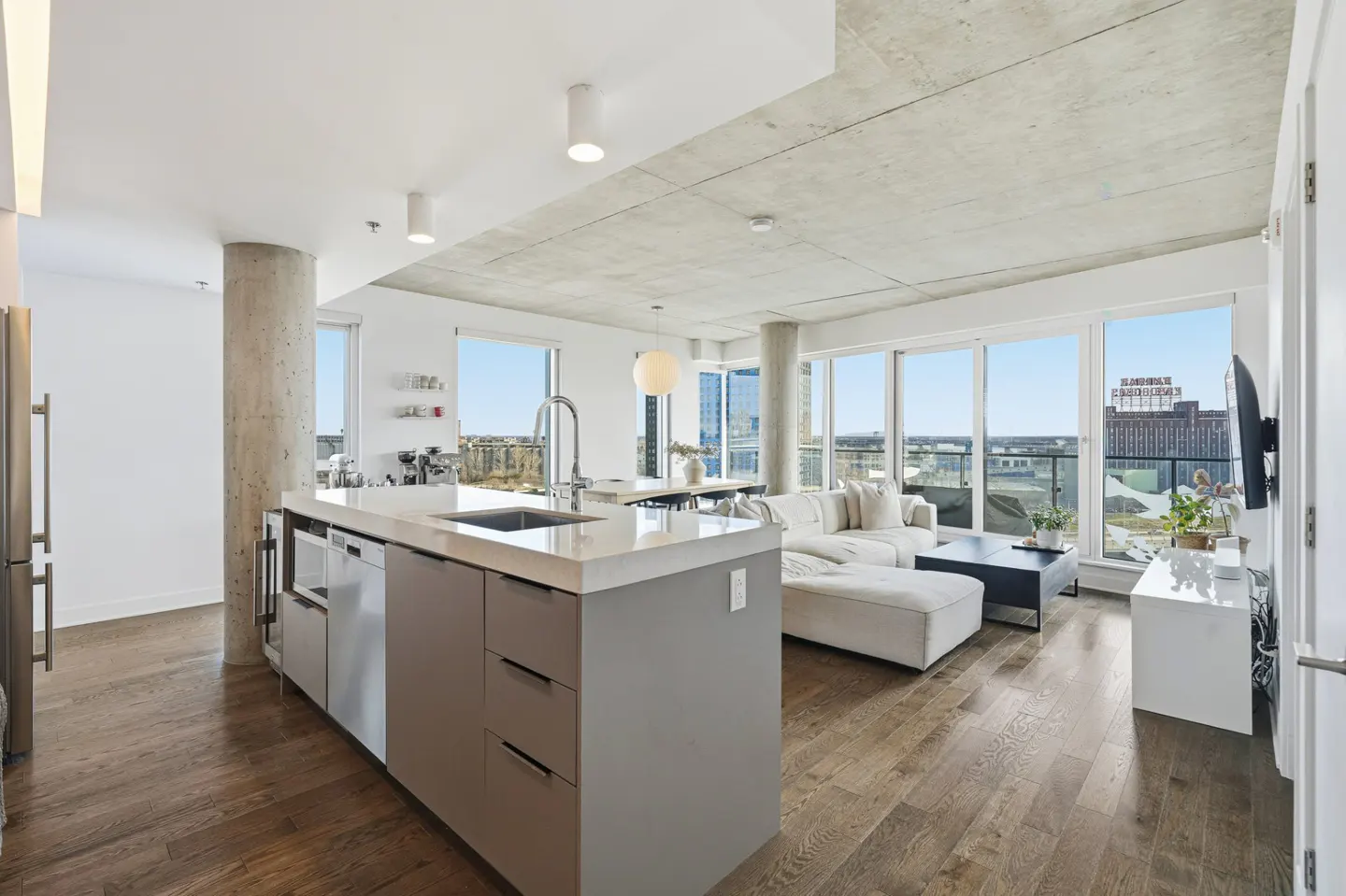 Open-concept condo with a kitchen island, stainless steel appliances, and a white sectional sofa near large windows with a city view.