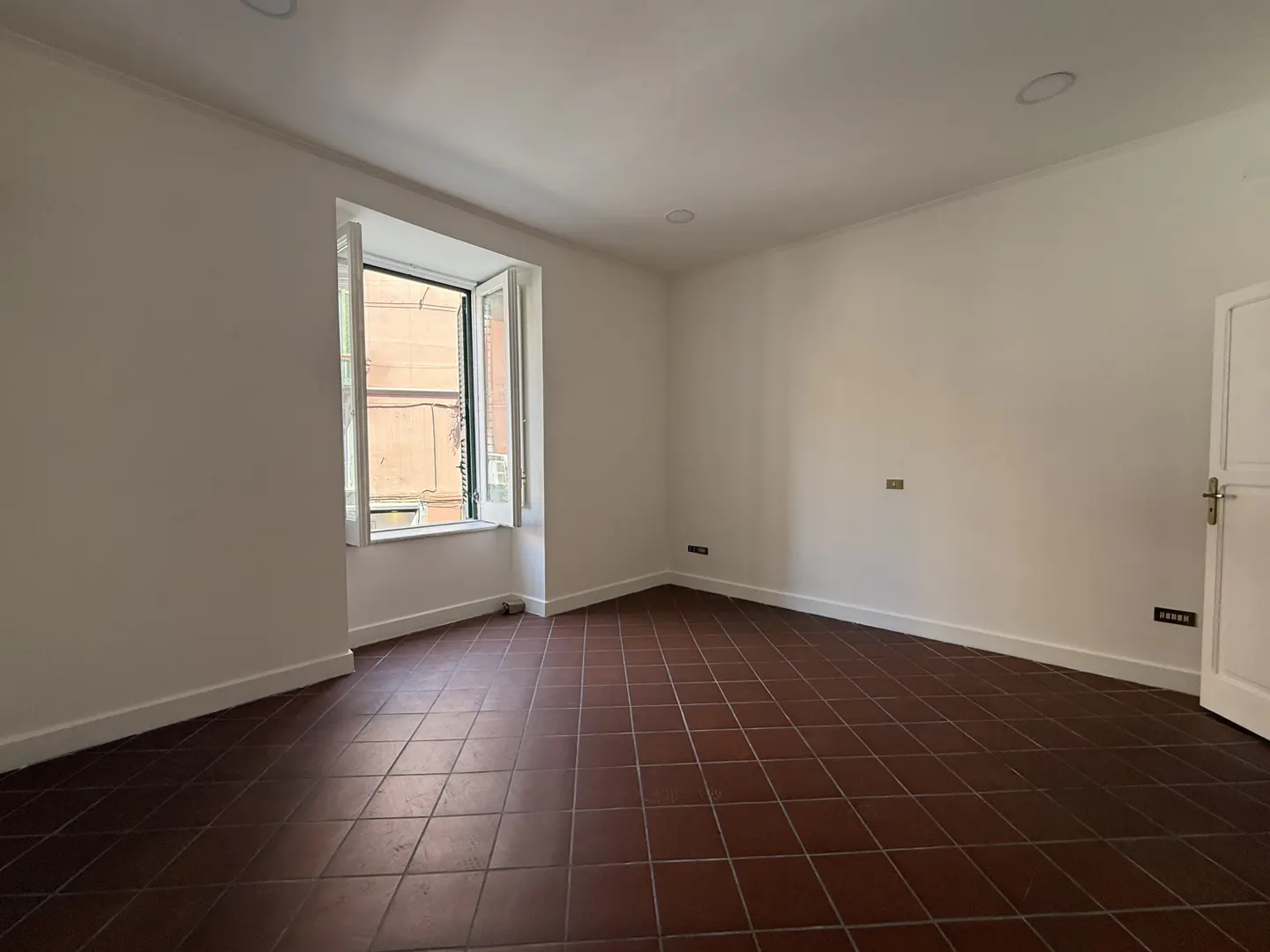 Bright, empty room with white walls, brown tiled floor, and an open window showing a building exterior.