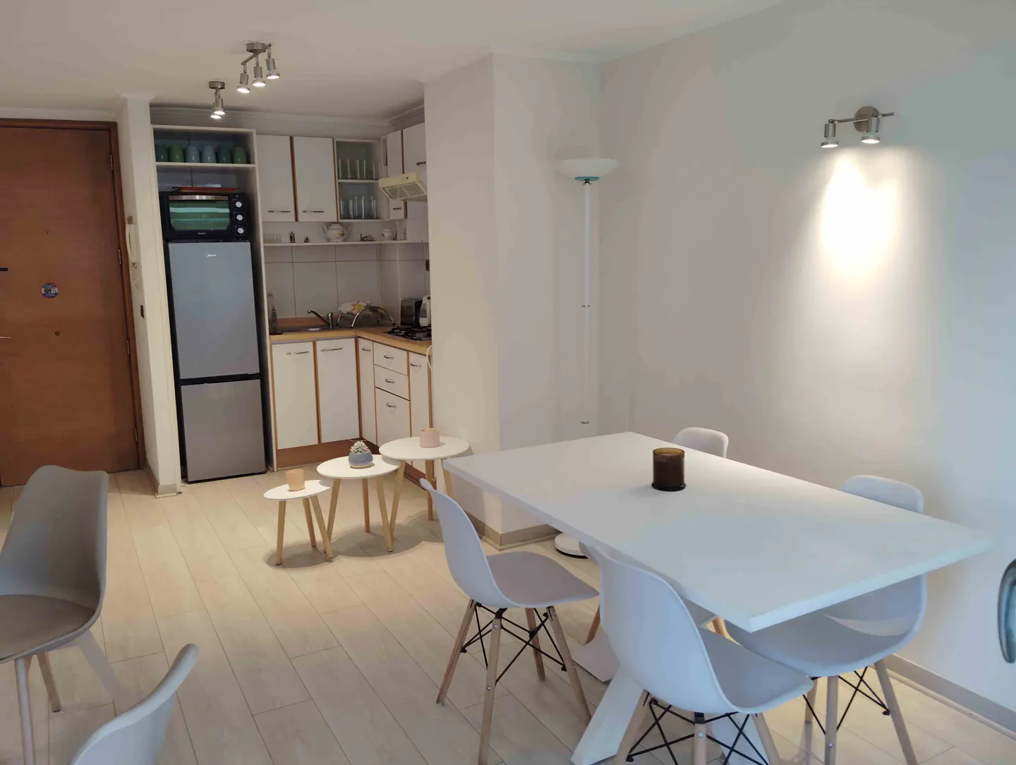 A bright, modern apartment interior with a white dining table and chairs. The kitchen features white cabinets and a stainless steel refrigerator.