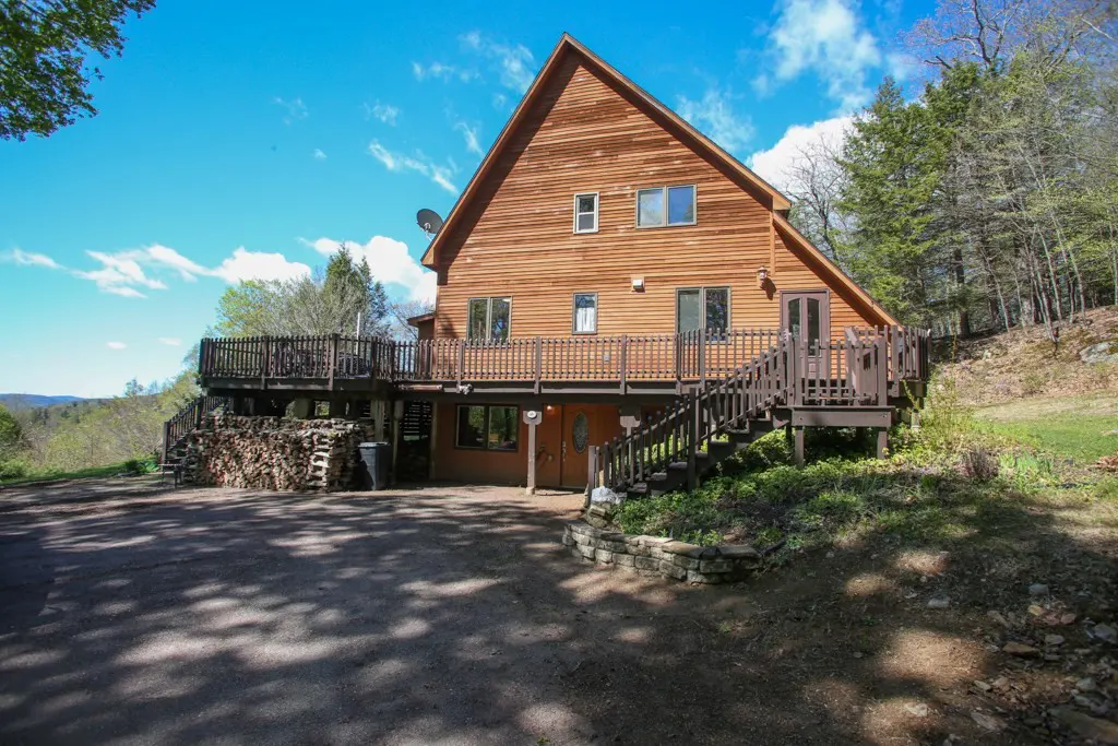 A two-story wood cabin with a deck and stairs, surrounded by trees and a blue sky.