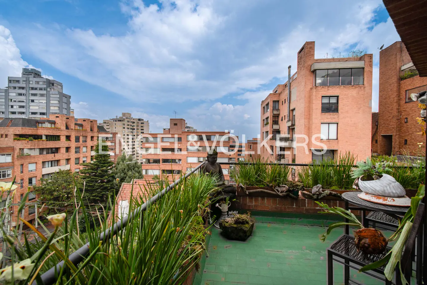 A green rooftop terrace with plants, furniture, and a statue, overlooking brick buildings under a cloudy sky.