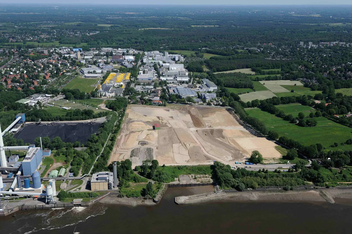 Aerial view of a construction site with a river in the foreground and a town in the background.