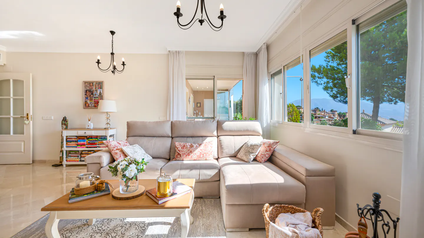 Bright living room with beige sectional sofa, wood coffee table, and large windows with mountain views. Chandeliers and white curtains add elegance.