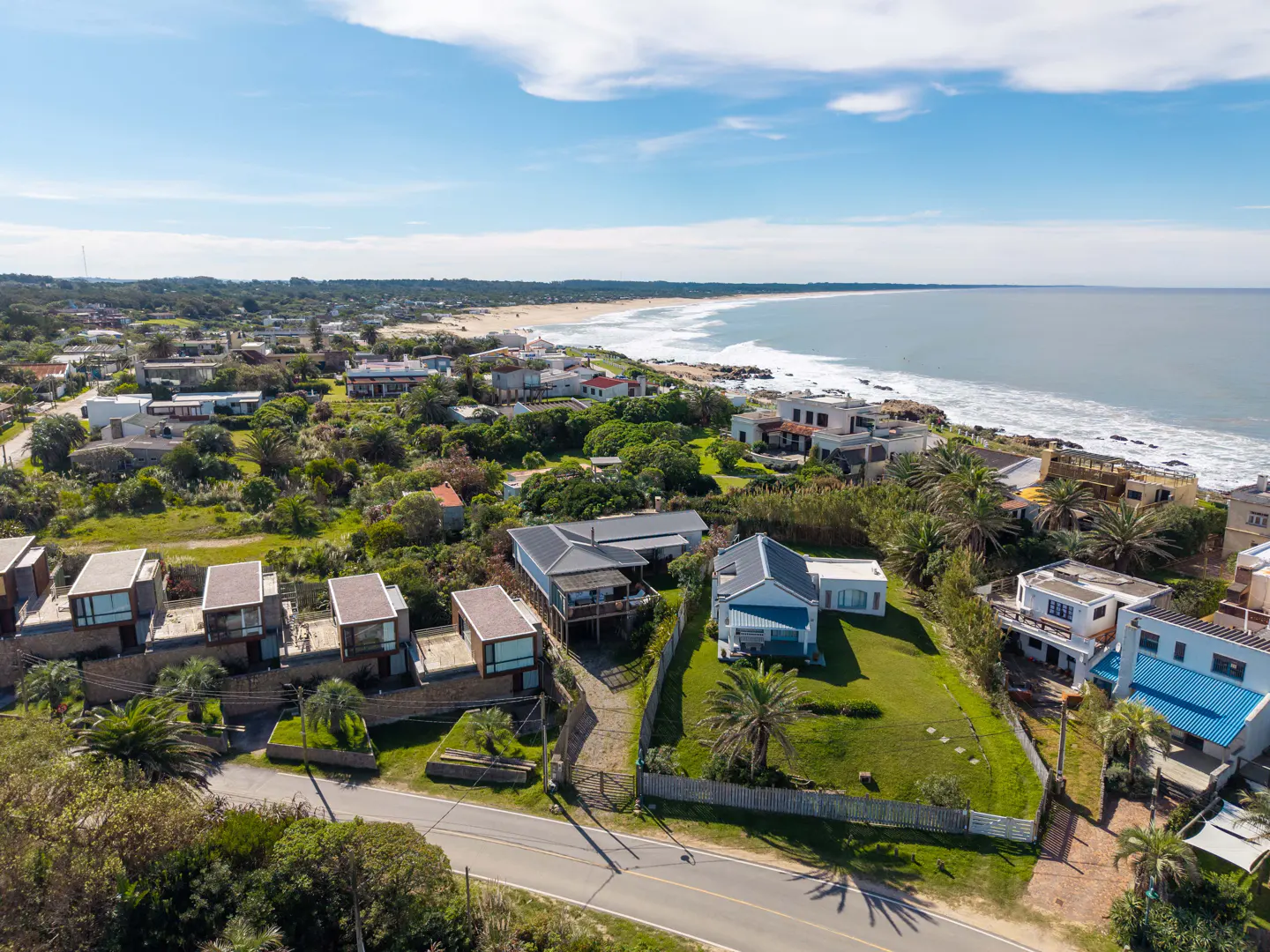 Aerial view of coastal homes with blue sky. Beachfront property with ocean waves and green landscape.