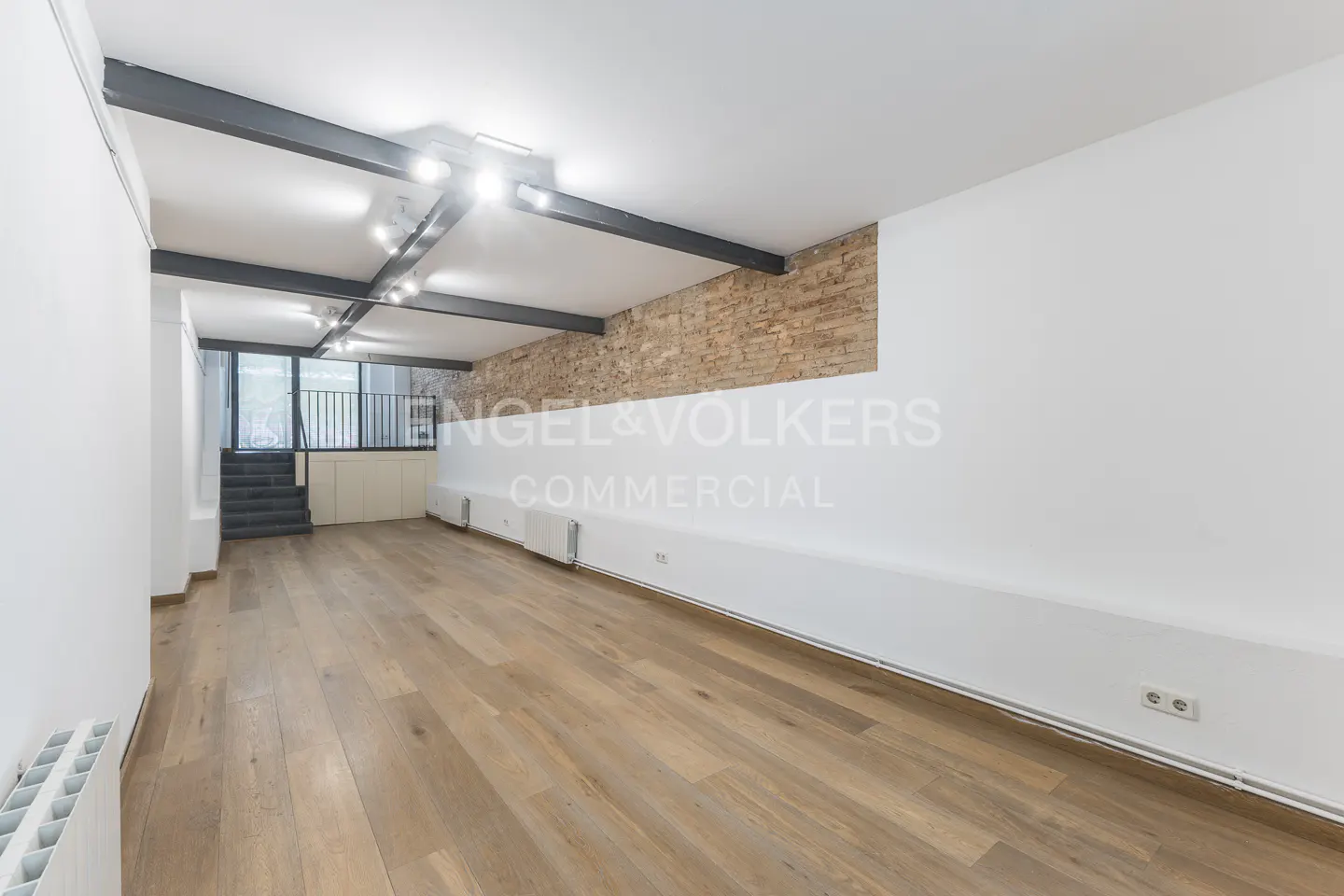 Bright, empty commercial space with hardwood floors, white walls, and exposed brick. Black beams cross the ceiling with track lighting. Stairs lead to a balcony.