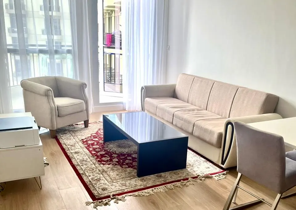 Bright living room with beige sofa and armchair, black coffee table on a red patterned rug, and large windows with sheer white curtains.