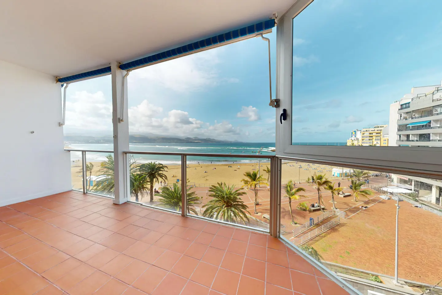 Balcony view of a sandy beach with palm trees, ocean, and buildings. The balcony has terracotta tiles and a glass railing.