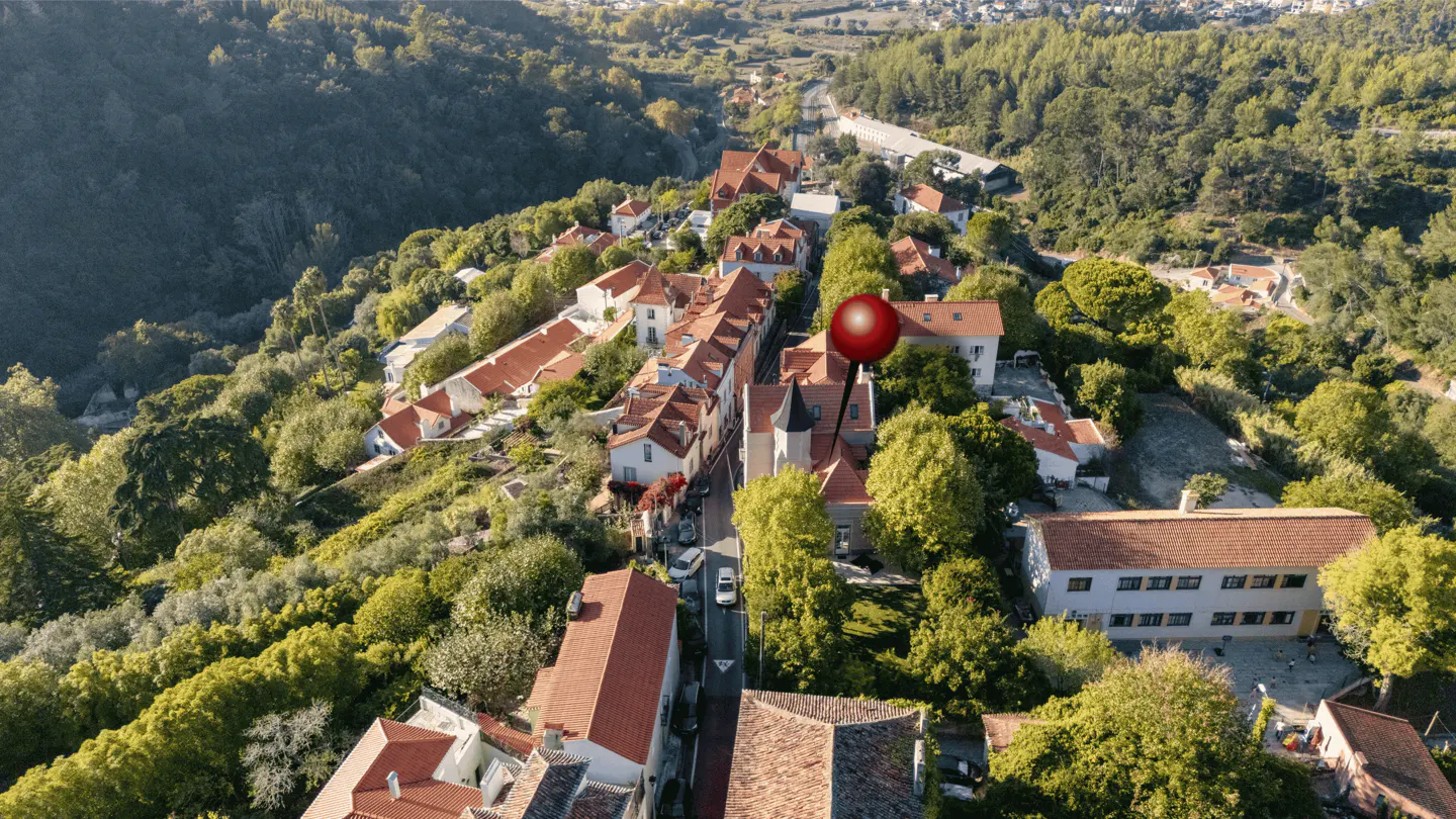 Aerial view of a European village with red-tiled roofs, surrounded by green trees and hills. A red pin marks a property.