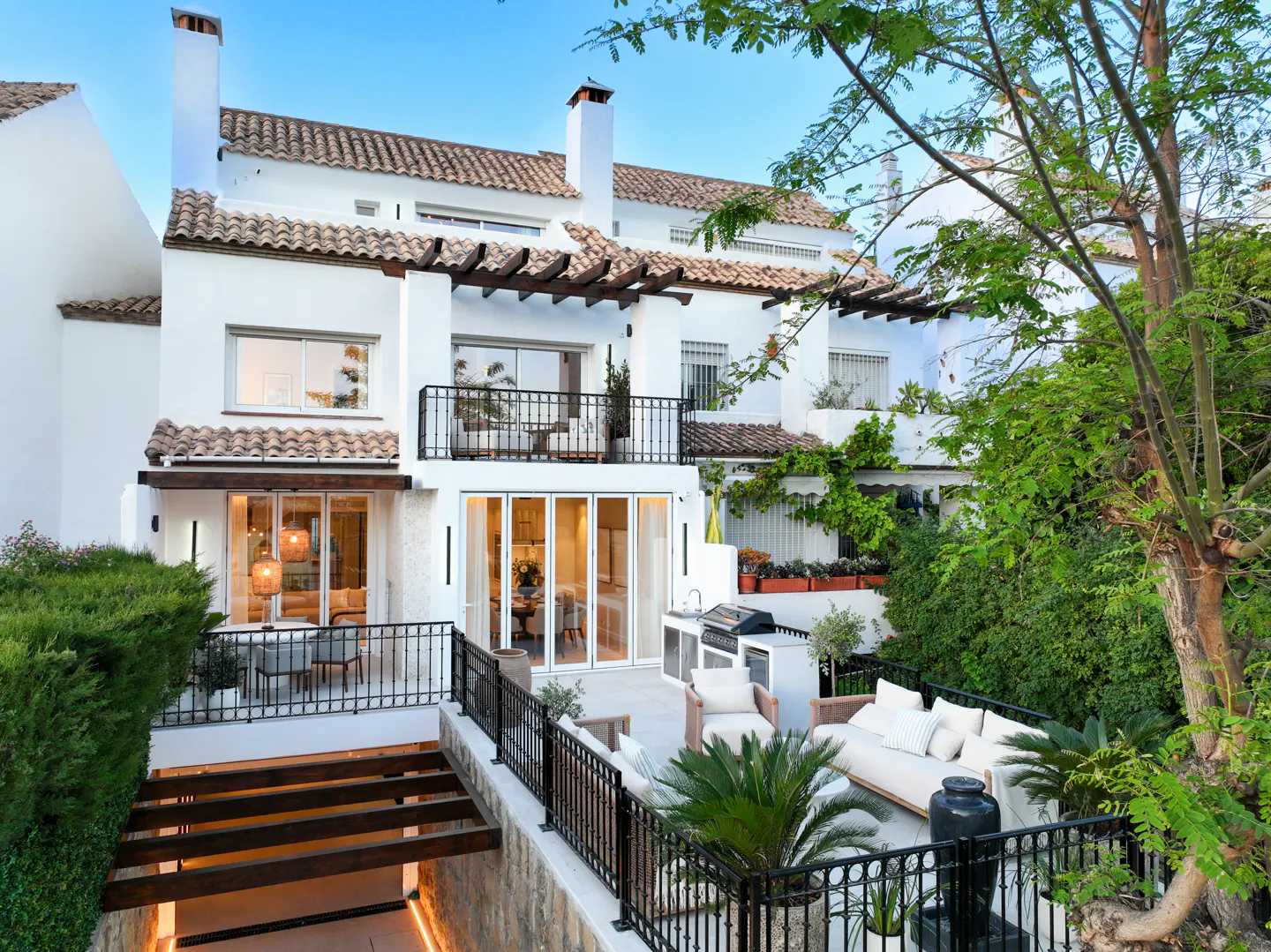 Exterior view of a white, multi-story house with a tile roof and black iron railings. A patio with outdoor furniture and a grill is visible. Lush greenery surrounds the property.
