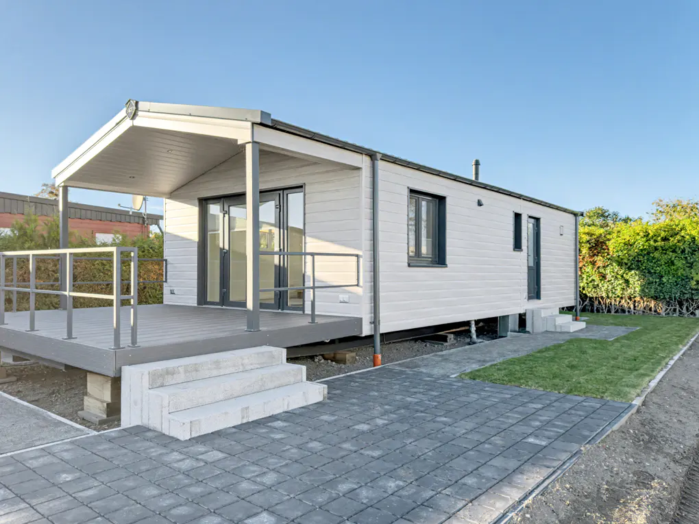 Exterior view of a white, modern mobile home with a gray deck, steps, and paved walkway. Blue sky background.