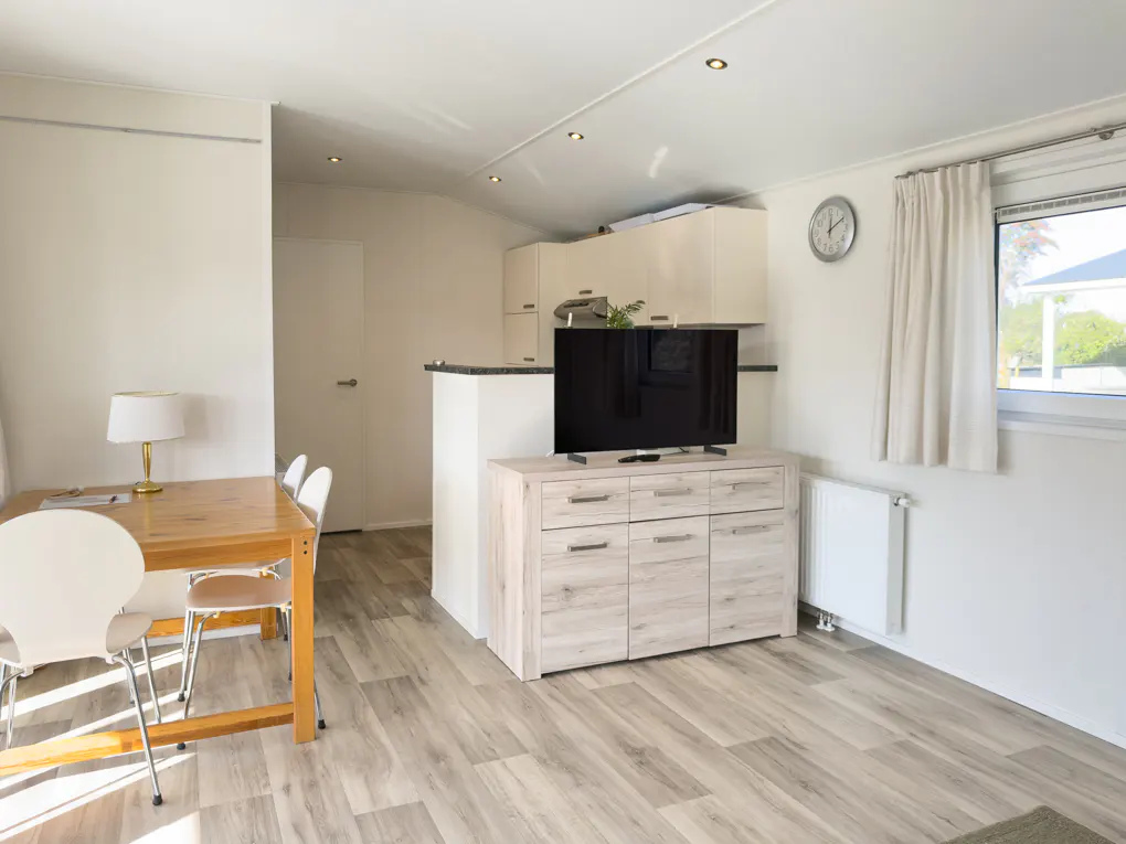 Bright living space with wood-look floors, a dining table with white chairs, and a TV on a light wood cabinet. Kitchen in the background.