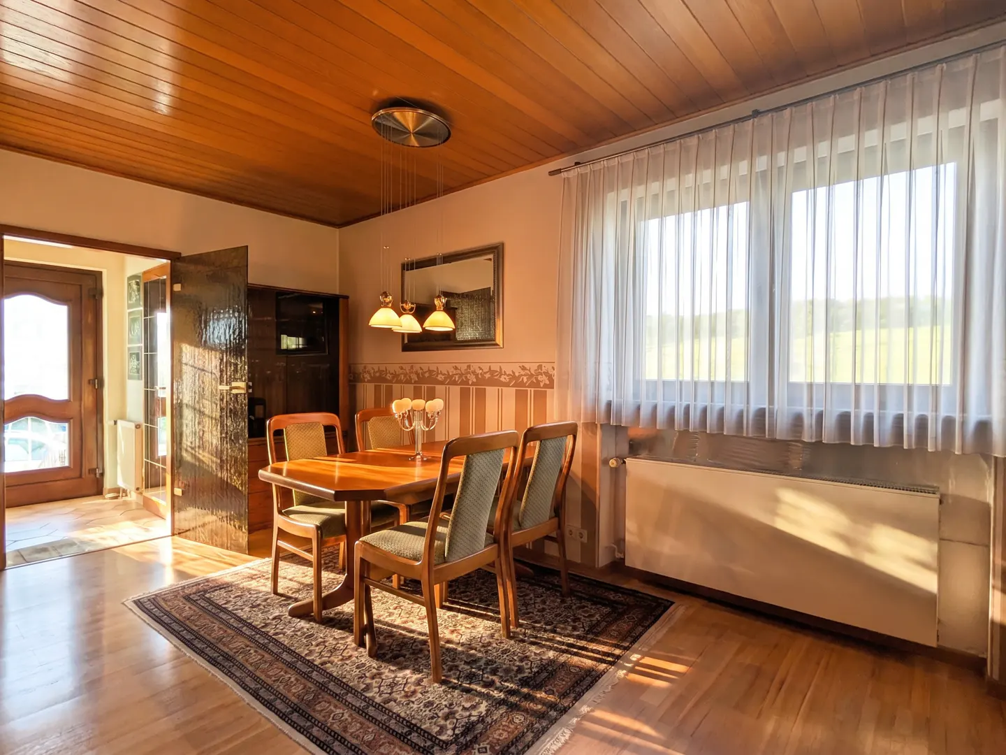 A dining room with a wood table, chairs, and rug. Sunlight streams through the window with sheer curtains.