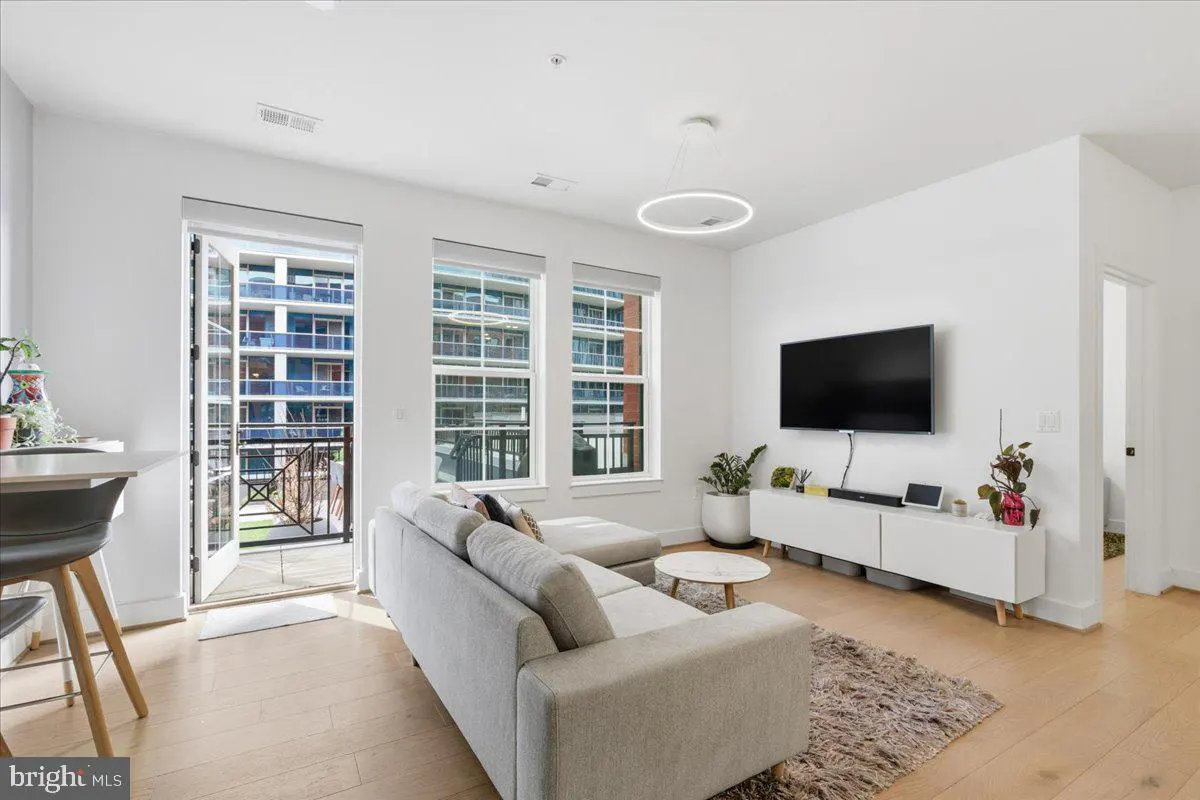 Bright living room with a gray sectional sofa, white walls, wood floors, and a balcony with a view of a blue building.